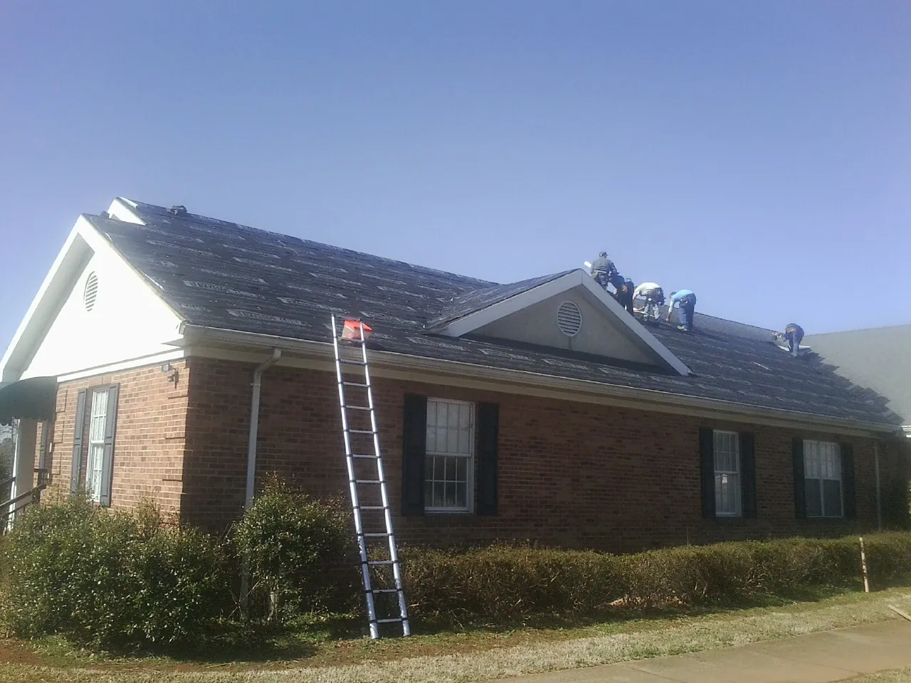Crew tearing shingles off a brick ranch roof with a ladder set — Metal Roof Repair work by our crew in Saint Michael