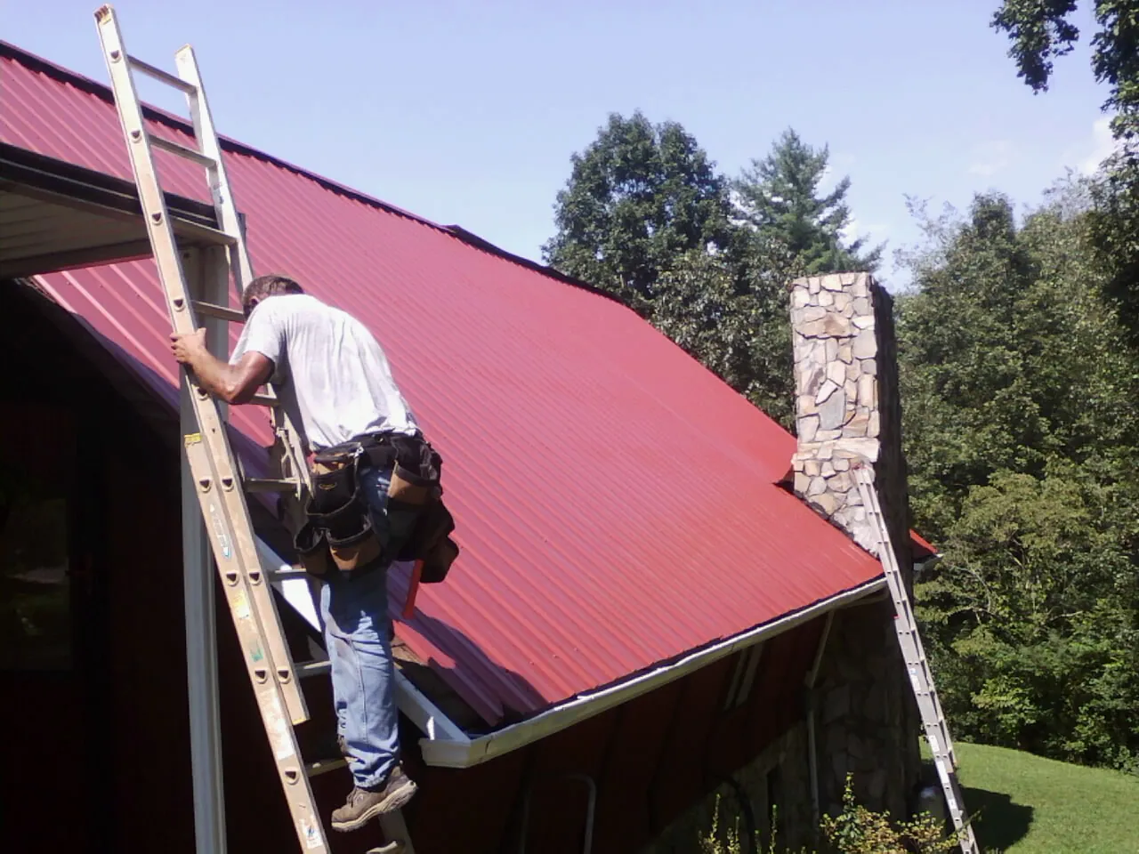 Roofer on a ladder beside a red metal roof and stone chimney — Soffit & Fascia Repair by Roofing Brothers in Saint Michael